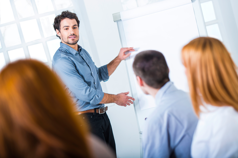 A male tutor in casual attire stands at a flipchart in front of a group of financial services students.