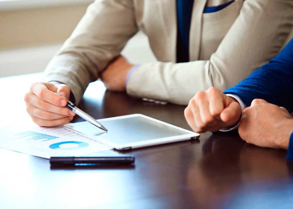 Two men study a financial advice report on a tablet. More financial paperwork lies underneath it.