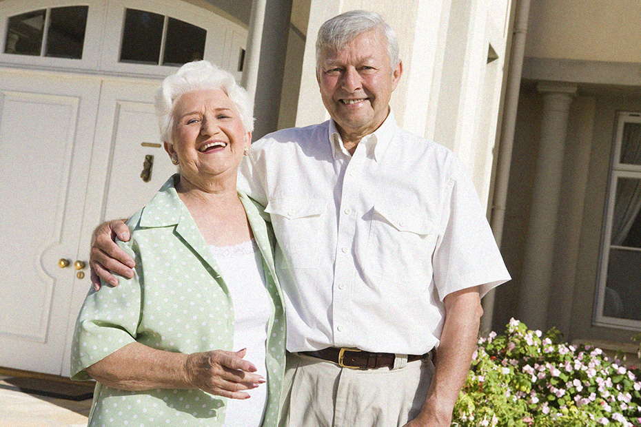 A senior man and woman standing in front of their house, they are casually dressed and smiling. Equity Release concept image.