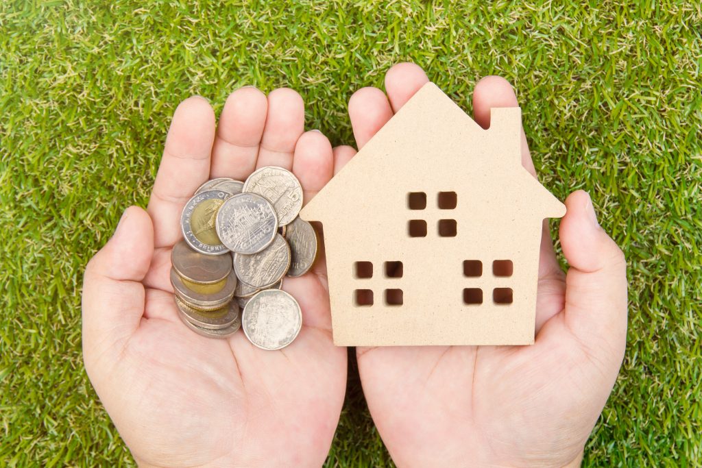 Mortgage concept image. A pair of hands hold a small wooden house and a pile of coins, set against a background of grass.