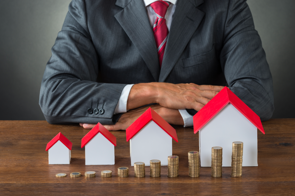 A male mortgage adviser in a grey suit sits in front of a row of model houses with a stack of coins in front of each house.