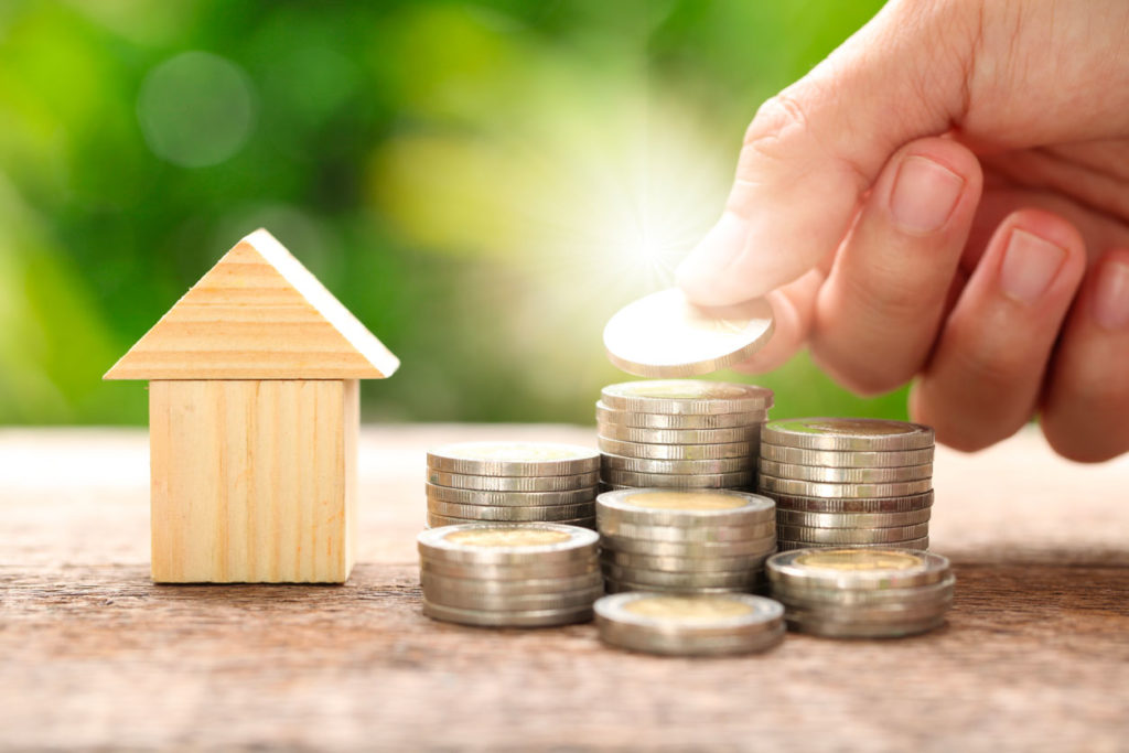 Mortgage advice concept. Stacks of coins stand next to a small wooden house. A man's hand adds a coin to one stack.