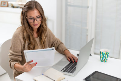 A female Independent Financial adviser uses a laptop as she checks financial services paperwork in a work-from-home setting.