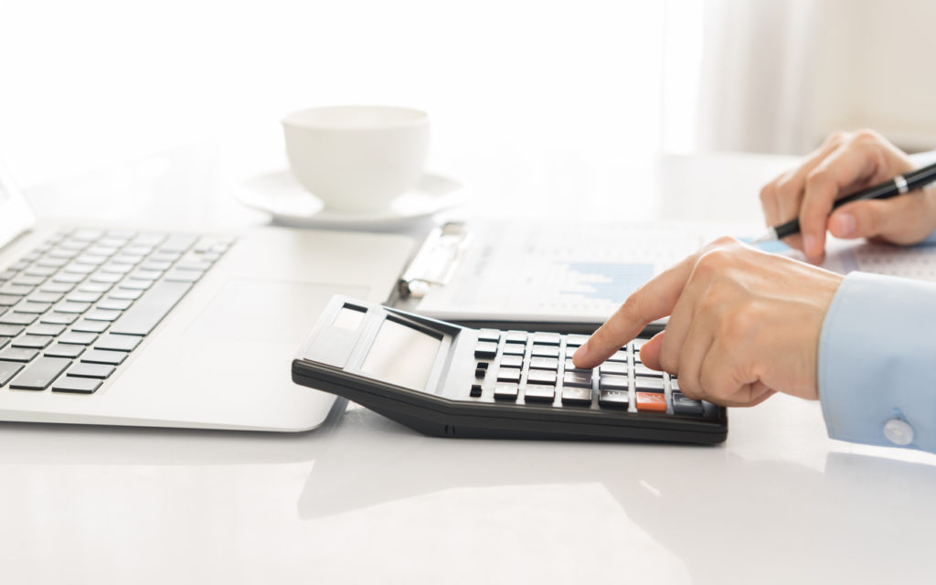 A man's hands using a calculator to work out figures for a financial advice report. A laptop and white cup are in the background.