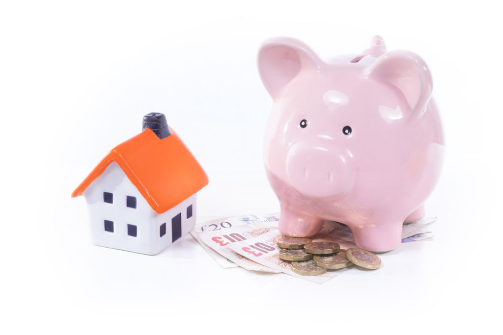 Mortgage advice concept. A pink china pig savings bank stands next to a small model house and a pile of coins and notes.