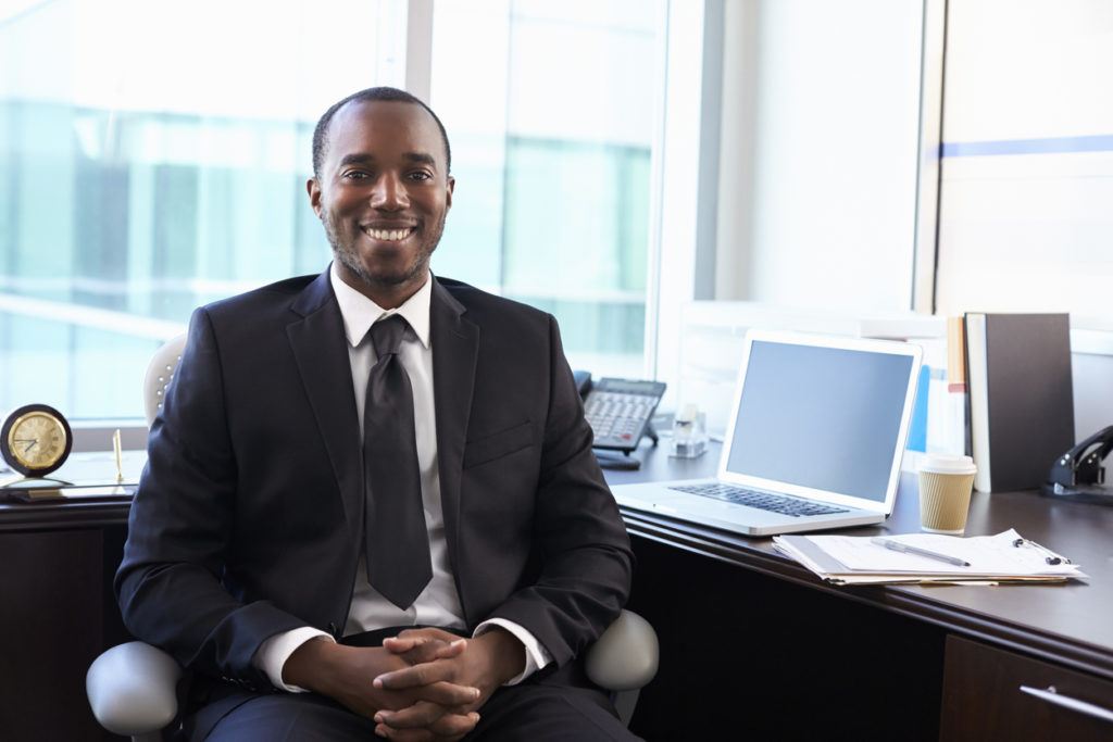 A Black male financial adviser sits in front of a desk in an office. He is wearing a suit and tie, and smiling confidently.
