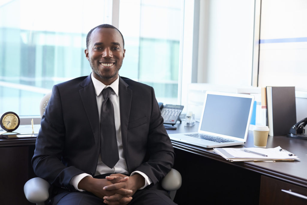 A Black male financial adviser sits in front of a desk in an office. He is wearing a suit and tie, and smiling confidently.