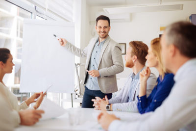 A male tutor stands at a whiteboard in front of a group of financial services students. He is smiling and holding a pen.