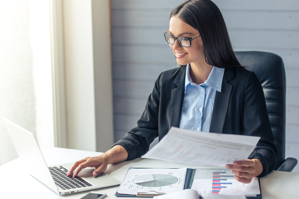 A young female financial adviser uses a laptop as she examines financial advice reports. She is smartly dressed and smiling.