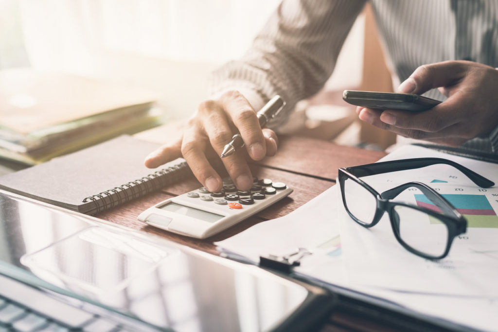 A male financial adviser is using a mobile phone and calculator while checking through financial advice reports.