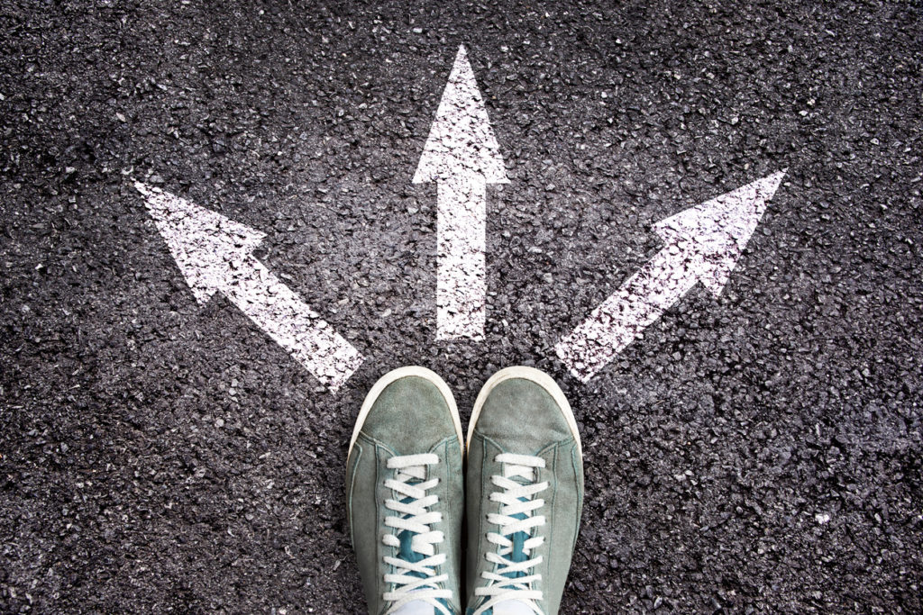 Decision concept image. 2 green trainers are shown in front of 3 arrows painted on tarmac, pointing in different directions.