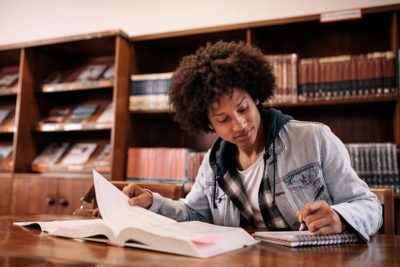 A young Black Financial Services student works at a table in a library. Shelves full of textbooks are in the background.