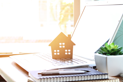 Mortgage advice concept. A small wooden house sits on a laptop keyboard, with sunlight shining through the window behind.