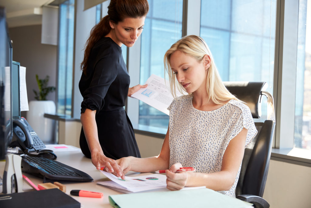 Two female financial advisers examine financial documents. One is sitting at an office desk and the other stands next to her.