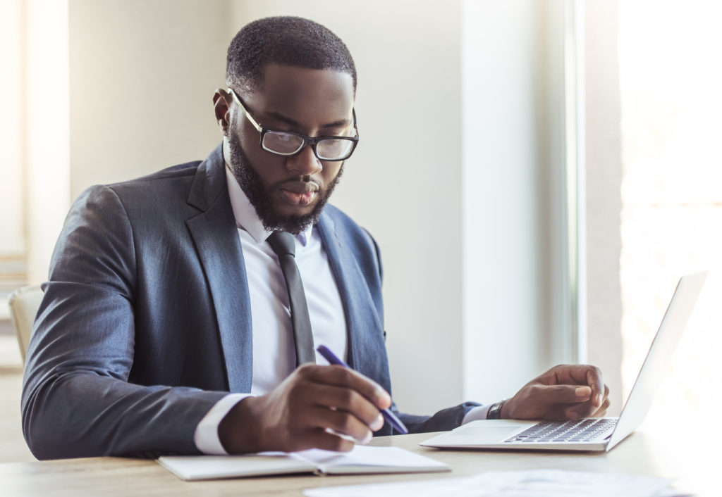 A Black male financial adviser studies his laptop while writing on a notepad. He is smartly dressed and focused on his work.