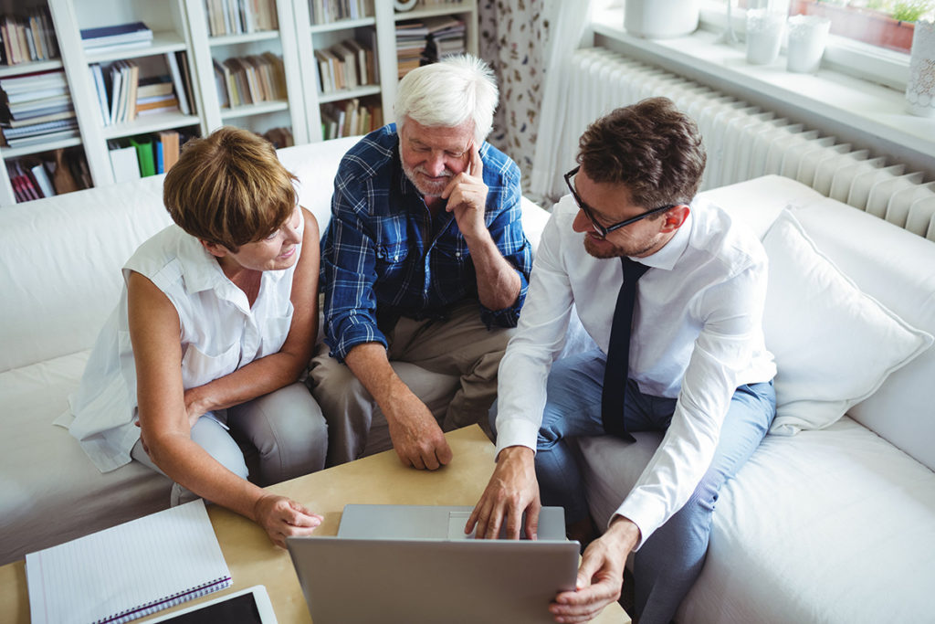A male Mortgage Adviser offers Equity Release advice to an elderly man and woman. They are looking at a laptop and smiling.