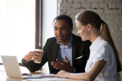 A man and woman in business clothes look at a laptop screen over cups of coffee