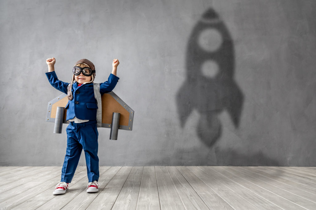 Young boy in a blue suit and cardboard wings with his hands in the air, creating a rocket ship shaped shadow