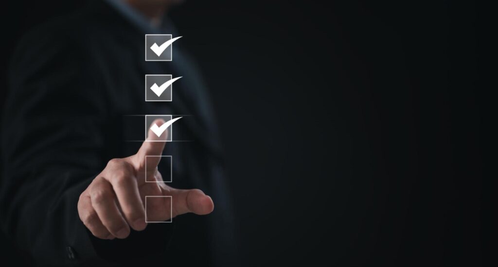 A person in a dark suit is pointing at a virtual checklist with four boxes, three of which are checked, representing task completion or progress tracking on a black background.