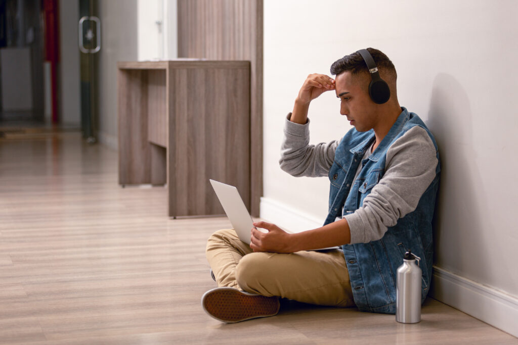 Person sitting on the floor with headphones, using a laptop, looking stressed.
