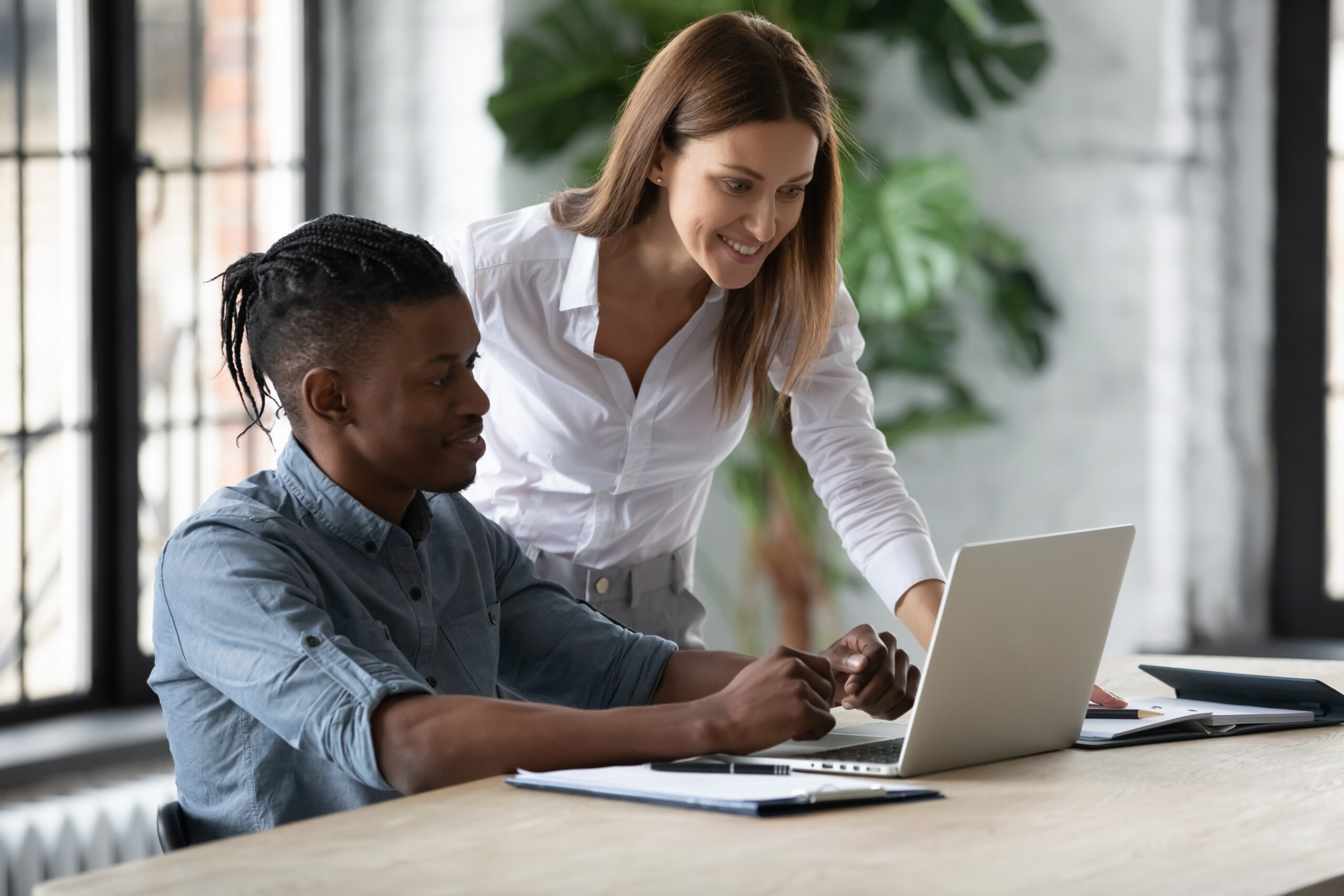 A mentor standing beside an apprentice, pointing at a laptop screen while guiding him through a task in a bright office workspace.